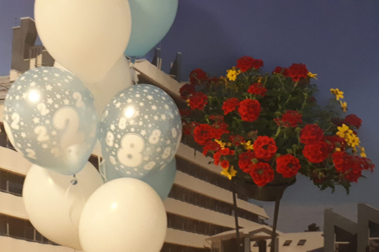 Zili un baltu balonu kopa ar 2 un 8 uz modernas ēkas fona. Blakus tiem piekārts grozs pārplūst ar košiem sarkaniem un dzelteniem ziediem. Debesis ir skaidras un zilas.