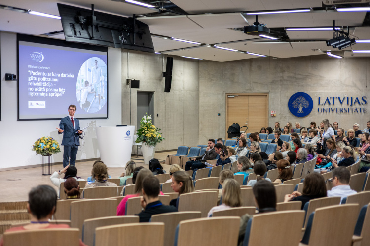 Konferenču telpa, krēslos sēž daudz cilvēki