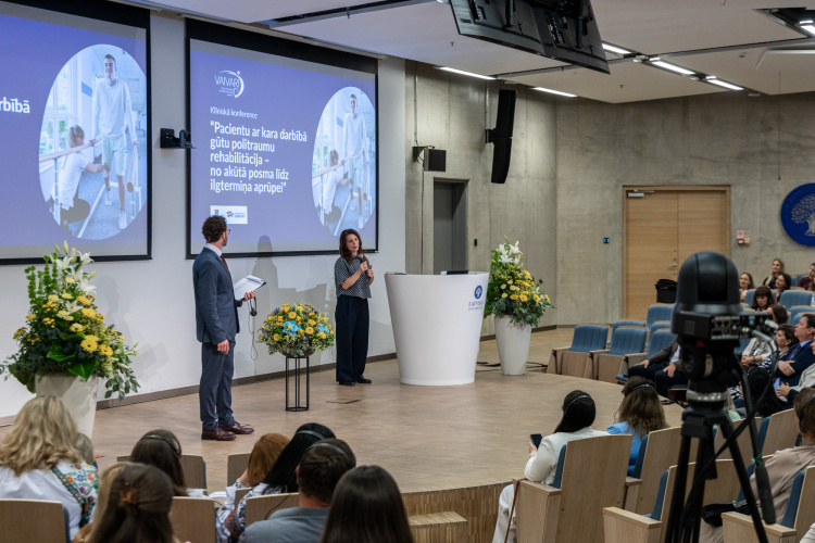 Lektors uzstājas uz skatuves modernā lekciju zālē, kurā auditorija sēž sēdvietās. Uz lieliem ekrāniem tiek rādīti medicīniski attēli un teksts. Telpu rotā ziedu kompozīcijas, un notikumu ieraksta kamera.