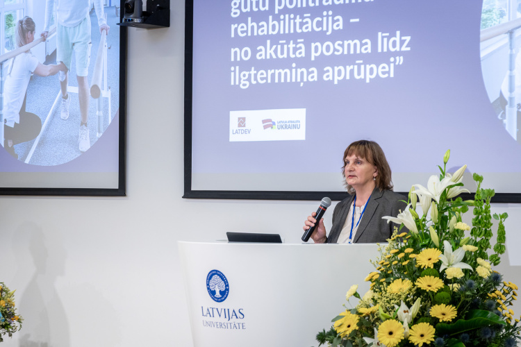 Sieviete runā pie tribīnes ar mikrofonu Latvijas Universitātē; priekšplānā ir ziedi, aiz viņas uz ekrāna redzams teksts latviešu valodā par rehabilitāciju.