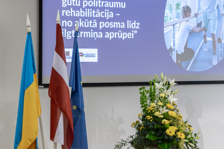 Konferenču zāle ar sēdošiem cilvēkiem, trīs karogiem (Ukrainas, Latvijas un ES), lielu ziedu pušķi un ekrānu, uz kura latviešu valodā tiek rādīta prezentācija par rehabilitāciju pēc kara traumām.