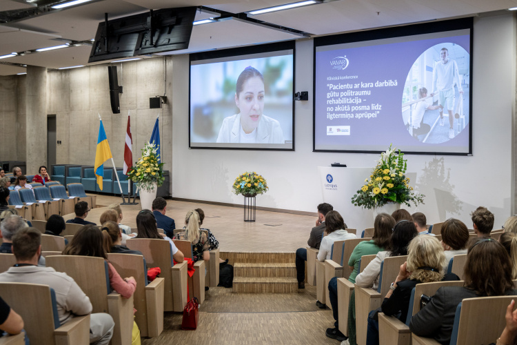 Konferenču zāle, kurā cilvēki sēž pretī skatuvei, kur uz diviem ekrāniem tiek rādīta virtuāli runājoša sieviete un informācija par pasākumu latviešu valodā. Pie skatuves redzami arī karogi un ziedu kompozīcijas.