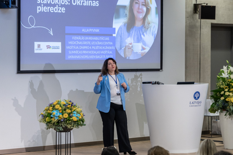 Sieviete zilā žaketē uzstājas uz skatuves un uzrunā auditoriju lekciju zālē. Aiz viņas ir liels ekrāns ar prezentācijas slaidu latviešu valodā un smaidošas sievietes fotogrāfiju. Skatuves laukumu rotā ziedi.