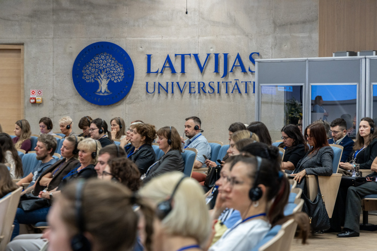 Cilvēku grupa sēž rindās, apmeklējot pasākumu Latvijas Universitātē. Daudzi ir ar austiņām un uzmanīgi klausās. Uz sienas aiz viņiem redzams universitātes logotips un nosaukums latviešu valodā.