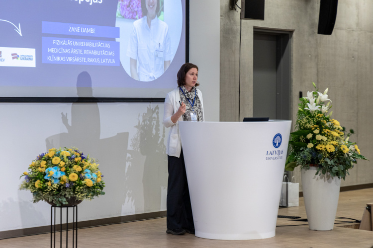 Sieviete stāv pie pjedestāla, uzstājoties ar prezentāciju modernā konferenču zālē, kurai abās pusēs ir ziedu kompozīcijas, aiz viņas ir liels ekrāns, bet uz pjedestāla - Latvijas Universitātes logotips.