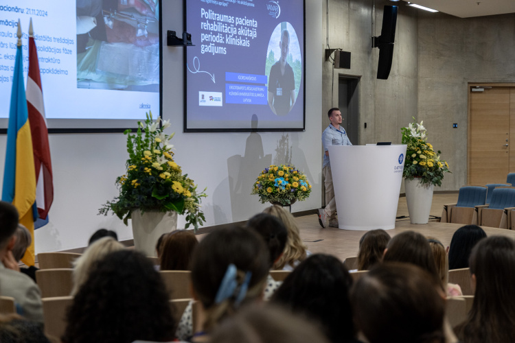 Vīrietis stāv pie pjedestāla un uzstājas ar prezentāciju modernā lekciju zālē, kur uz lieliem ekrāniem tiek rādīti diapozitīvi par pacientu rehabilitāciju. Klausītāji sēž, un skatuvi rotā ziedu kompozīcijas.