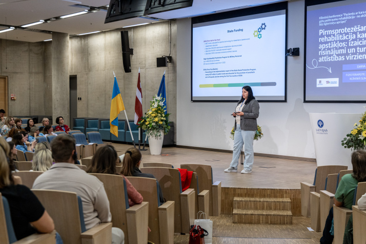 Uz skatuves stāv sieviete, kas uzstājas auditorijā un uzstājas ar prezentāciju. Aiz viņas ir liels ekrāns, uz kura tiek rādīti diapozitīvi, un pa kreisi redzami Ukrainas un Latvijas karogi.
