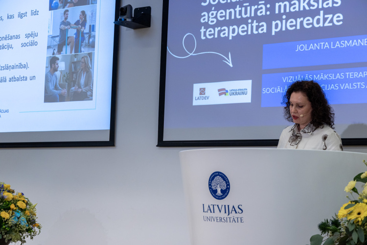 Sieviete stāv pie tribīnes ar uzrakstu Latvijas Universitate, uzstājoties pasākumā. Aiz viņas ir slaidi ar tekstiem un fotogrāfijām, vienā no tiem minēta Jolanta Lasmane. Uz skatuves redzamas ziedu kompozīcijas.