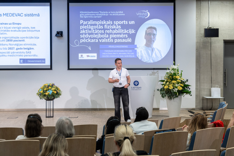 Uz skatuves konferencē uzstājas vīrietis, kurš runā uz skatuves pie projekcijas ekrāna, uz kura tiek rādīta informācija par paralimpiskajiem sporta veidiem un rehabilitāciju. Klausītāji sēž viņam pretī modernā lekciju zālē ar izstādītiem ziediem.