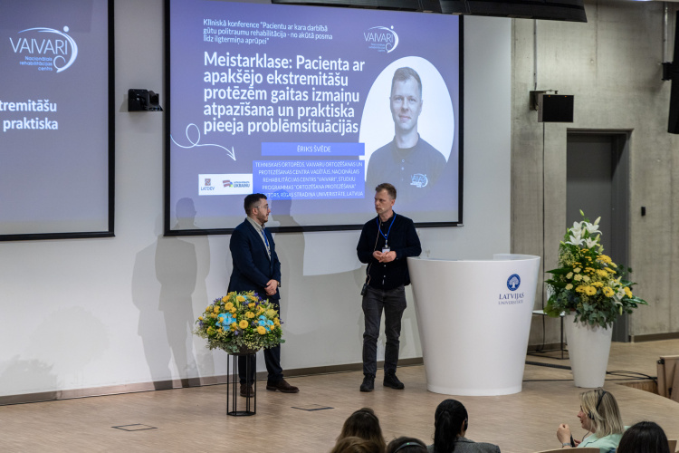 Divi vīrieši stāv uz skatuves blakus lektorijam un lieliem ekrāniem, uz kuriem tiek rādīta prezentācija latviešu valodā. Izskatās, ka notiek konference, priekšplānā redzamas puķu kompozīcijas un auditorija.