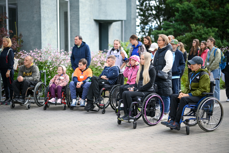 Cilvēku grupa, tostarp vairāki cilvēki ratiņkrēslos, pulcējas ārā uz bruģēta laukuma. Daži no viņiem sēž, bet citi stāv aiz viņiem, un šķiet, ka visi uzmanīgi klausās vai vēro kaut ko ārpus kadra.