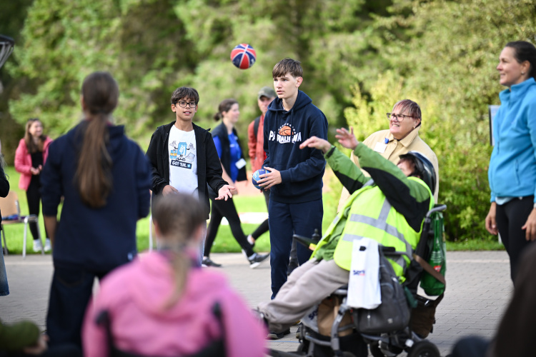 Grupa cilvēku, tostarp bērni un cilvēks ratiņkrēslā, spēlē ar basketbola bumbu ārā uz bruģēta laukuma apstādījumu ieskautā laukumā. Visi izskatās aizņemti un smaidīgi.