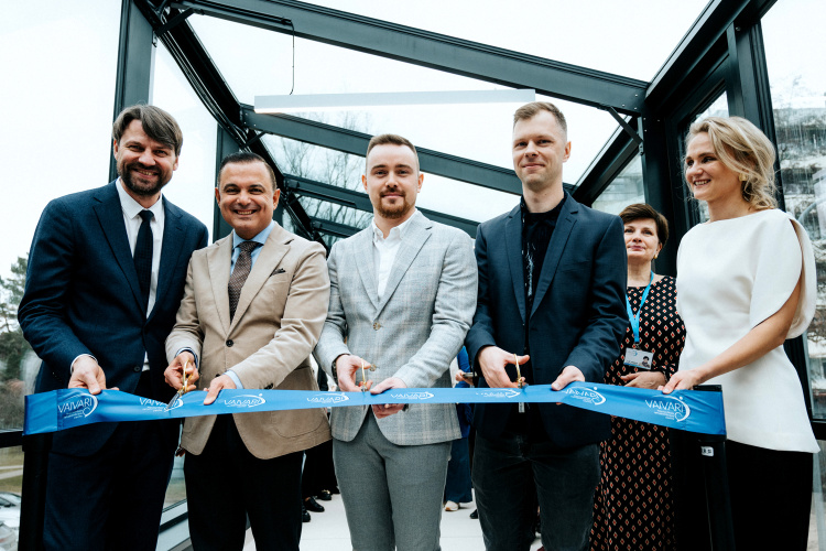 Seši cilvēki, tērpušies svinīgā tērpā, smaida un tur rokās šķēres, griežot zilu lentīti svinīgā lentes griešanas ceremonijā iekštelpās. Pasākums šķiet svinīgs, un fonā redzamas stikla sienas un pilsētas skats.