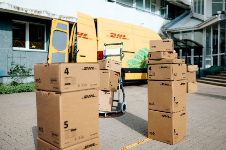 Pie ēkas ieejas uz zemes priekšā dzeltenajam DHL piegādes mikroautobusam ir sakrautas vairākas DHL kartona kastes, un blakus atrodas ratiņi.