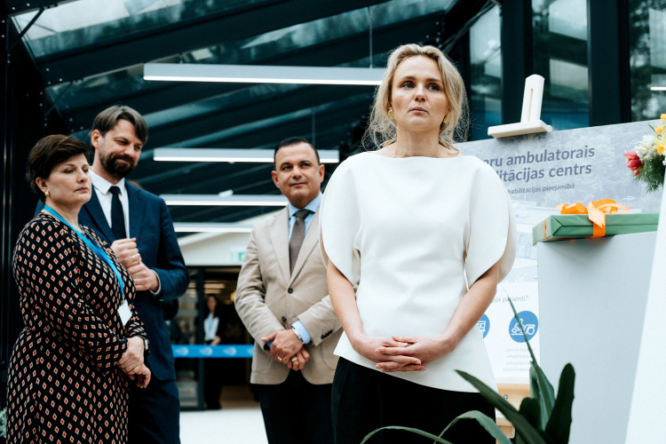 Četru profesionāli tērptu cilvēku grupa stāv iekštelpās pie pjedestāla ar lenti, izskatoties uzmanīgi un nopietni kāda pasākuma vai ceremonijas laikā. Fonā redzama moderna stikla arhitektūra.