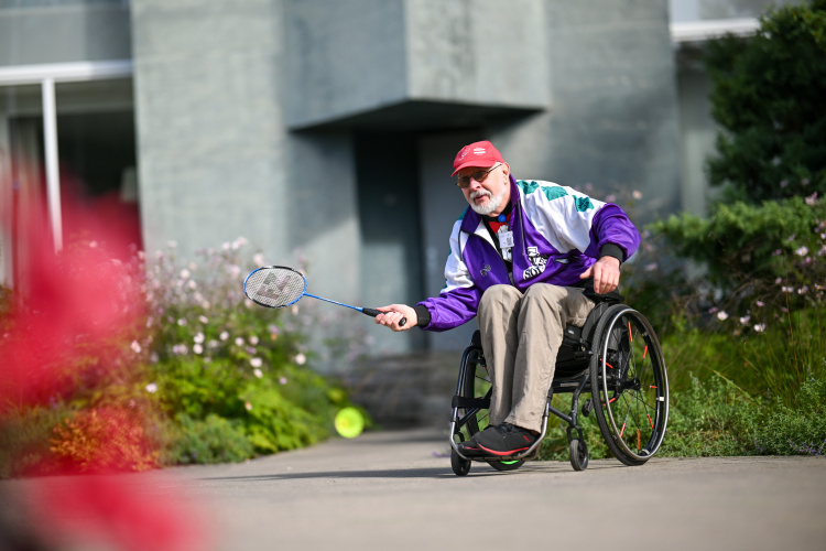 Vecāks vīrietis ratiņkrēslā, sarkanā cepurē un violetā jakā, spēlē badmintonu ārā uz bruģēta celiņa, fonā ir zaļumi un ziedi.