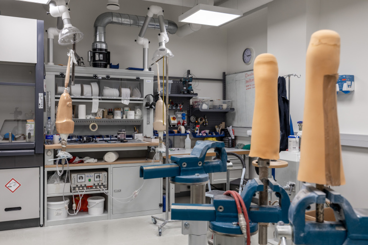 Laboratorijas darba telpa ar protezētām ekstremitātēm skavās, instrumentiem, plauktiem ar aprīkojumu un dažādiem materiāliem, kas izkārtoti pa visu telpu.