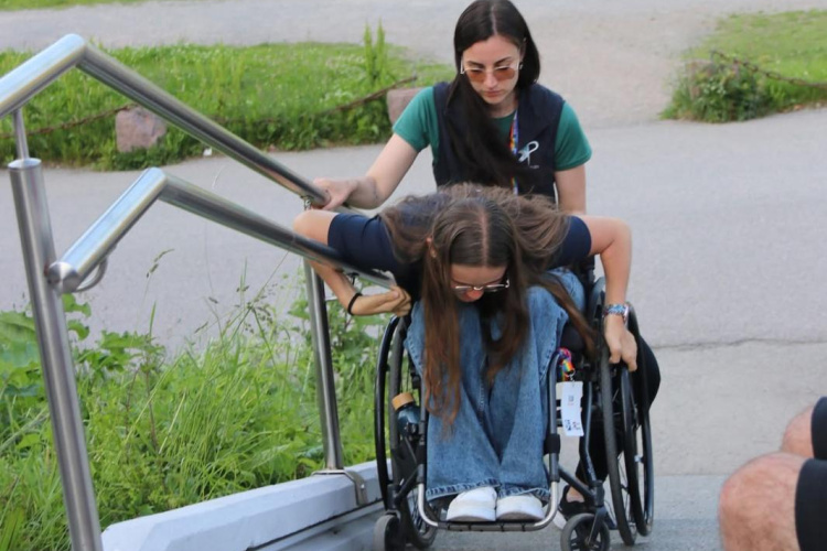 Sievietei ratiņkrēslā palīdz cita sieviete, kad viņas mēģina uzbraukt pa apstādījumu ieskautiem āra kāpnēm. Asistente tur rokturus, palīdzot ratiņkrēslam virzīties augšup.