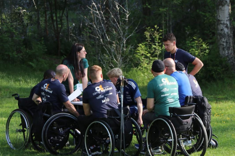 Cilvēku grupa ratiņkrēslos pulcējas aplī uz zaļās zāles saulainā parkā ārā un iesaistās sarunās, fonā redzami koki un zaļumi.