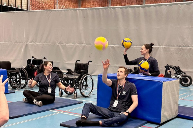 Trīs cilvēki sēž sporta zālē uz paklājiņiem, met un ķer dzeltenas volejbola bumbas. Fonā redzami ratiņkrēsli. Izskatās, ka vide ir sporta nodarbība vai terapijas nodarbība cilvēkiem ar kustību traucējumiem.