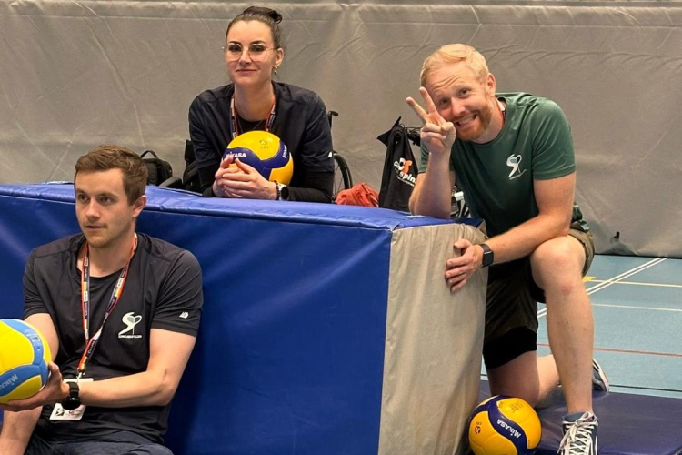 Trīs pieaugušie sēž uz paklājiņiem slēgtā sporta zālē ar volejbola bumbām; viens vīrietis smaida un mirkšķina ar miera zīmi, sieviete sēž aiz polsterētas barjeras un tur bumbu, bet otrs vīrietis sēž, sakrustojis kājas un arī tur bumbu.