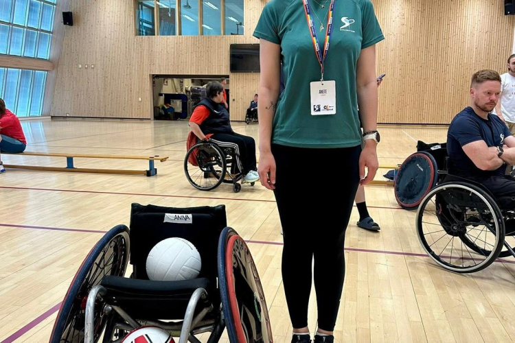 Sieviete zaļā kreklā ar jostiņu stāv sporta zālē blakus sporta ratiņkrēslam ar volejbola bumbām. Fonā uz koka grīdas redzami vairāki cilvēki ratiņkrēslos.