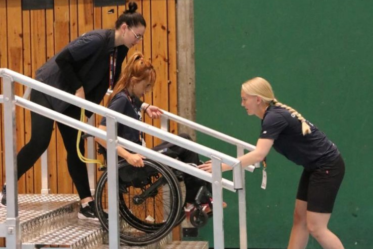 Cilvēku ratiņkrēslā pa metāla uzbrauktuvi pāri kāpnēm vada divi palīgi sporta zālē, viens stumjot no aizmugures, bet otrs velkot no priekšpuses.