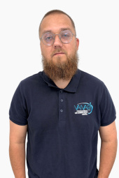 Vīrietis ar bārdu un brillēm, kurš valkā tumši zilu polo kreklu ar Vanari Neirozinātņu centra logotipu, stāv uz vienkārša balta fona.