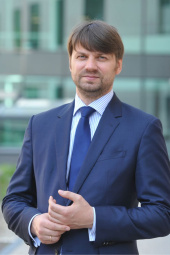 Vīrietis zilā uzvalkā un kaklasaitē stāv ārā pilsētvidē ar modernām stikla ēkām fonā un ar vieglu smaidu raugās uz kameru.