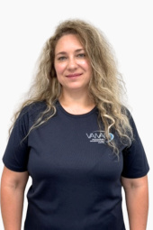 Sieviete ar gariem, cirtainiem blondiem matiem smaida un ir tērpusies tumši zilā T-kreklā ar nelielu „VANA“ logotipu kreisajā krūšu pusē; viņa stāv uz vienkārša balta fona.
