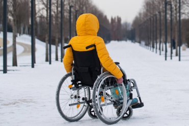 Cilvēks spilgti dzeltenā jakā sēž ratiņkrēslā uz sniegota celiņa, ko ieskauj kaili koki un laternu stabi, un ir vērsts prom no kameras.