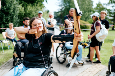 Saulainā dienā jauna sieviete ratiņkrēslā vērš loku un bultu uz āra lokšaušanas pasākumā, apjozta ar citiem dalībniekiem, no kuriem daži arī atrodas ratiņkrēslos.