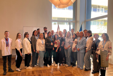 Cilvēku grupa, gan vīrieši, gan sievietes, stāv kopā labi apgaismotā telpā ar lieliem logiem, koka grīdām un modernu gaismekli, pozējot grupas fotoattēlam un smaidot kamerai.